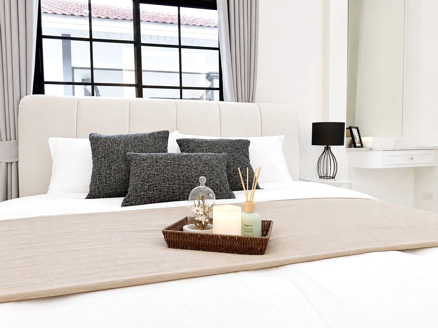 Neutral-toned bedroom with a white upholstered bed, dark gray pillows, and a beige runner; a wicker tray holds a candle and diffuser on the duvet.