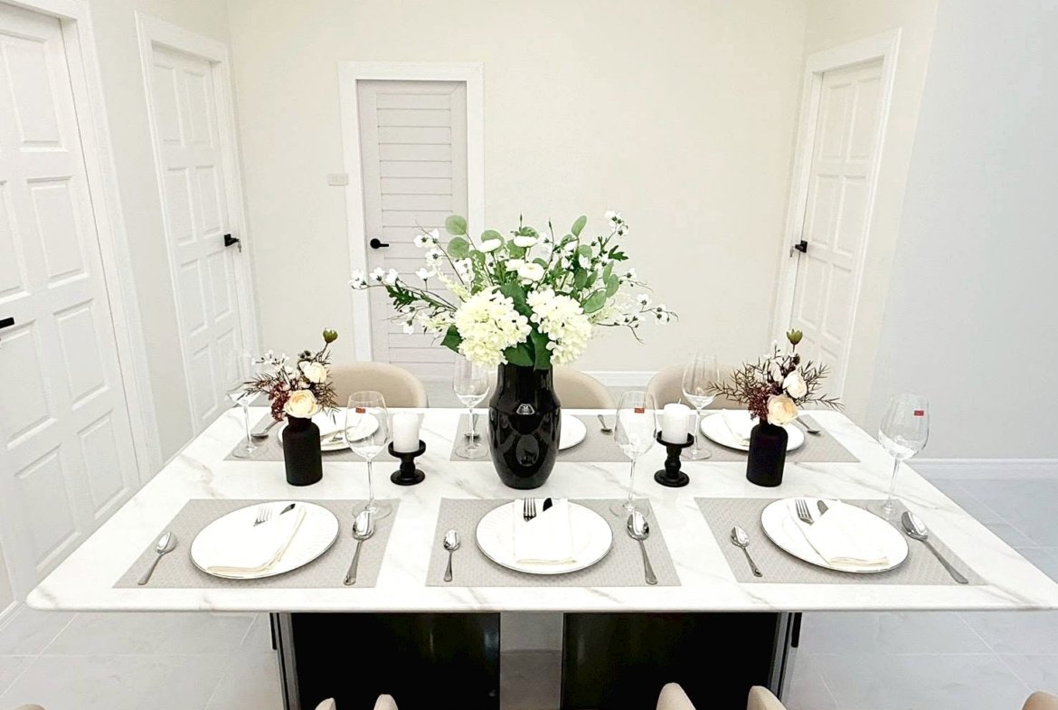 Formal dining table set for six with beige chairs, whitePlace settings, and a tall black vase centerpiece of white flowers.