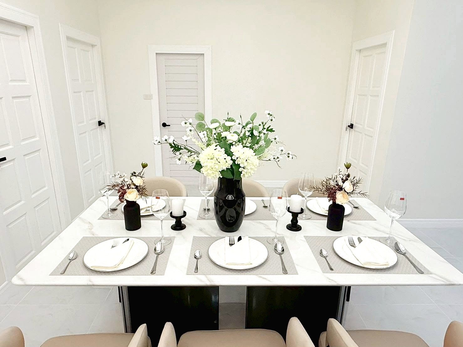 Formal dining table set for six with beige chairs, whitePlace settings, and a tall black vase centerpiece of white flowers.