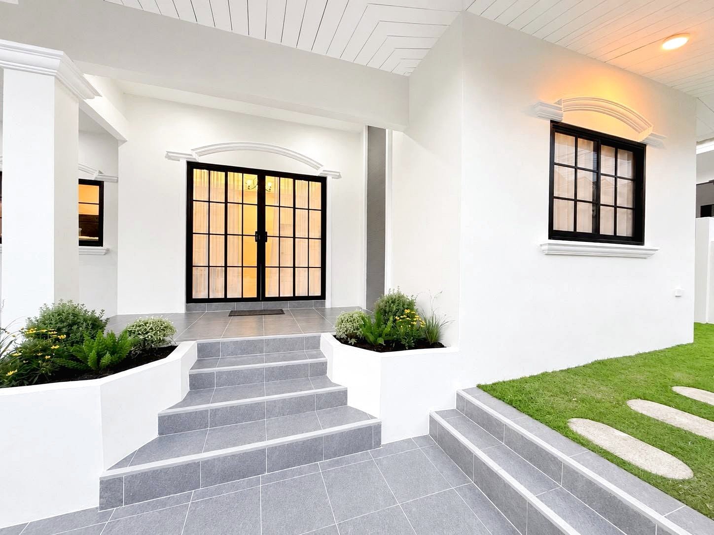 White modern house entry with gray tile steps and black-framed glass doors, surrounded by small garden beds.