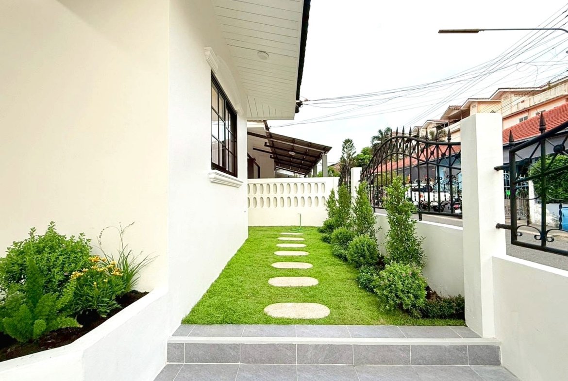 Narrow grassy path with circular stepping stones leading to a white wall gate, between a house and a black wrought-iron fence.