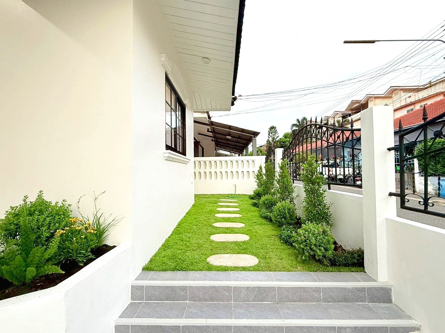Narrow grassy path with circular stepping stones leading to a white wall gate, between a house and a black wrought-iron fence.