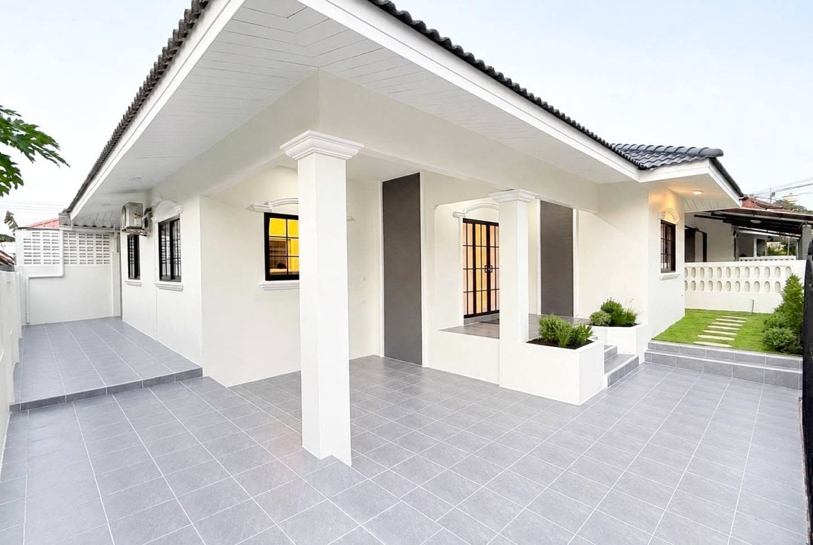 White single-story house with a wide tiled courtyard, supported by columns, and a small front garden area with planters and steps leading to the entrance