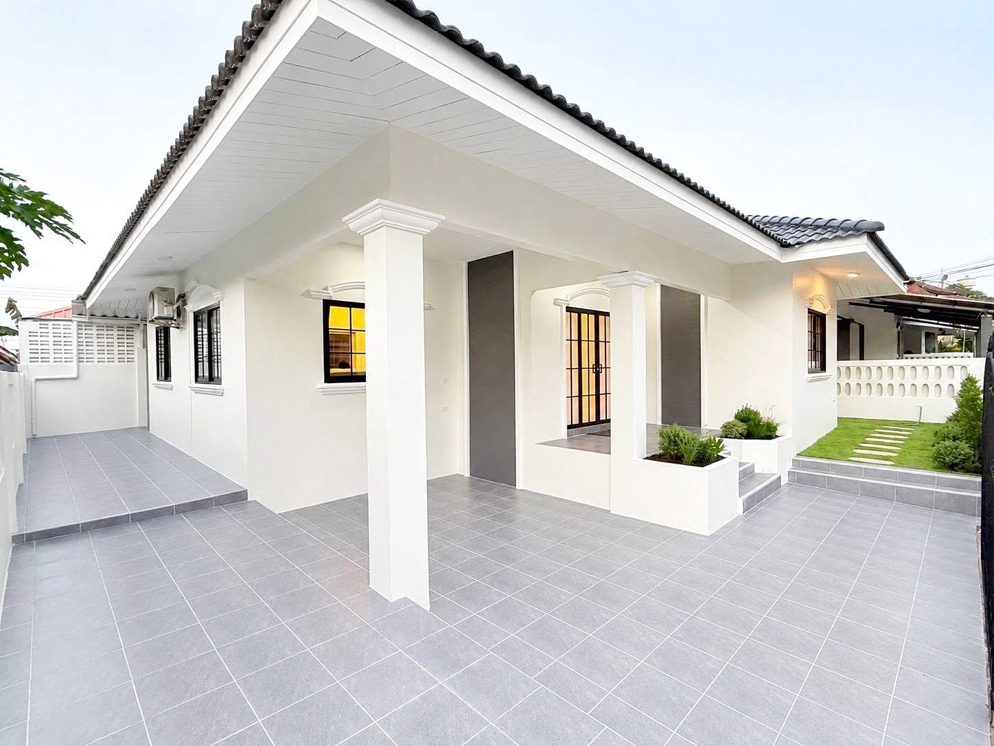 White single-story house with a wide tiled courtyard, supported by columns, and a small front garden area with planters and steps leading to the entrance