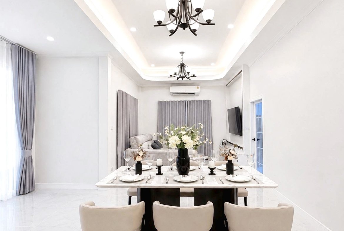 Bright dining room with a marble table set for eight, white china, candles, and a black vase of white flowers; living area with gray curtains behind.