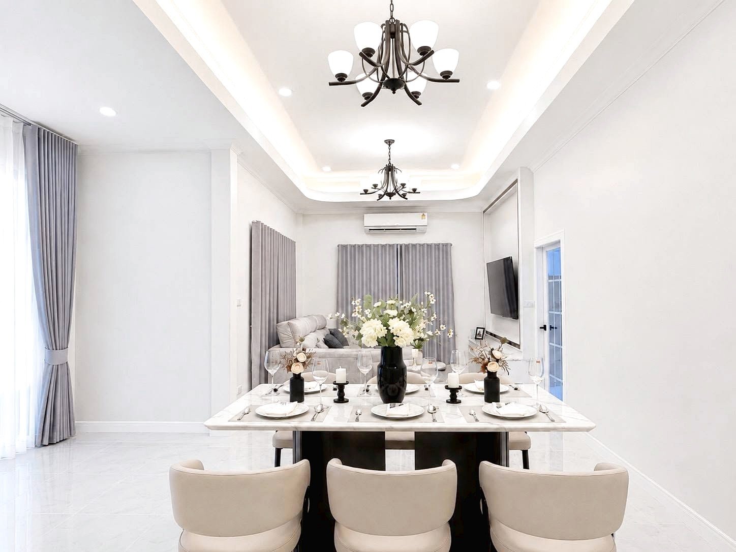 Bright dining room with a marble table set for eight, white china, candles, and a black vase of white flowers; living area with gray curtains behind.