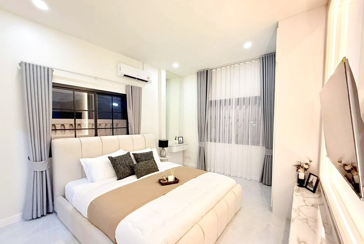 Bright modern bedroom with a large beige upholstered bed, tan runner, and gray throw pillows under soft lighting into a neutral palette.