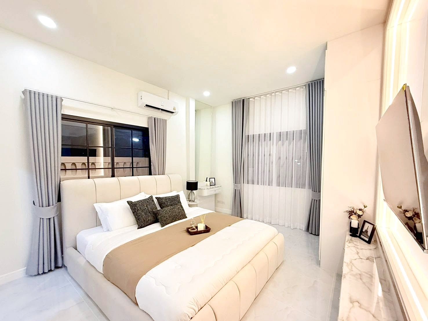 Bright modern bedroom with a large beige upholstered bed, tan runner, and gray throw pillows under soft lighting into a neutral palette.