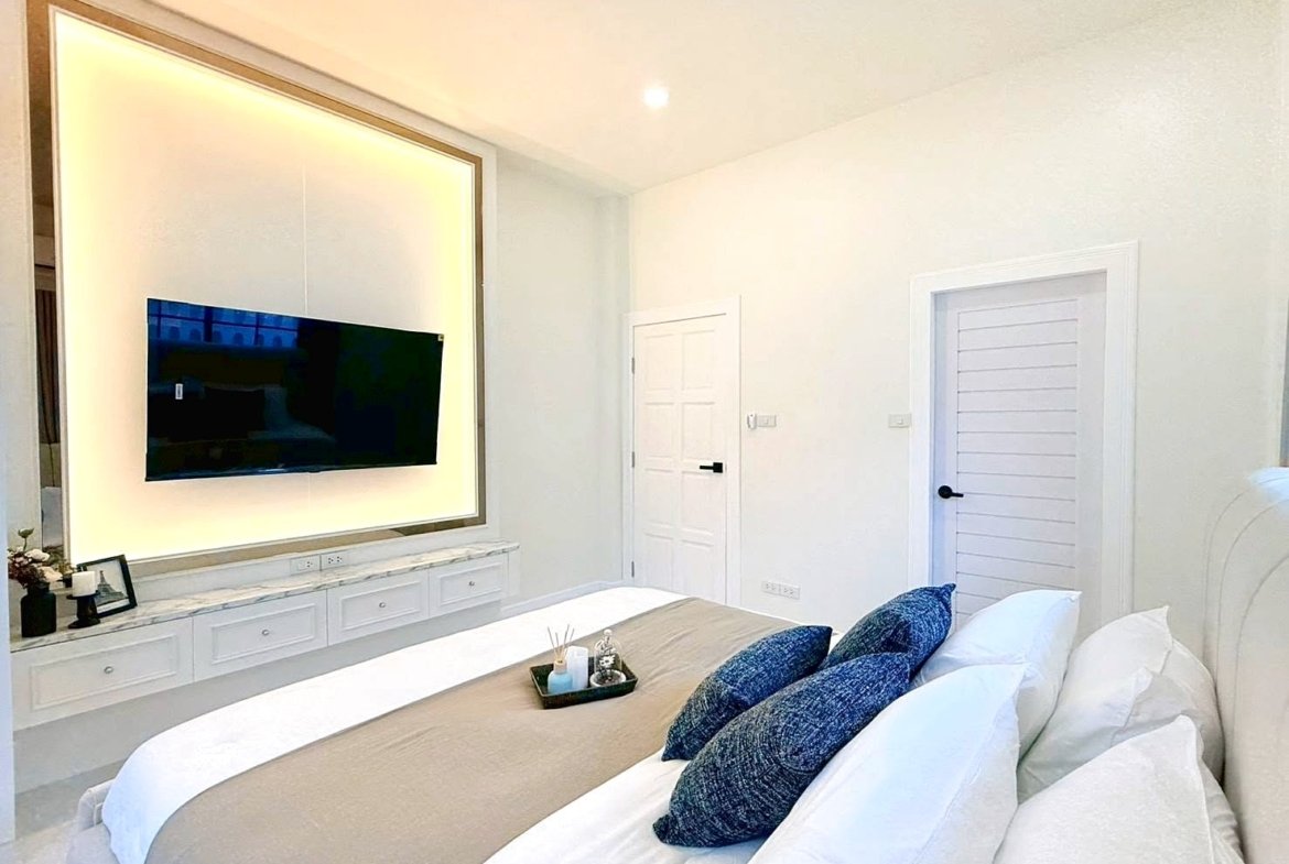 Bright modern bedroom with a wall-mounted flat-screen TV set in a glowing framed inset above a white marble console and drawers, overlooking a neatly made bed with blue accent pillows.
