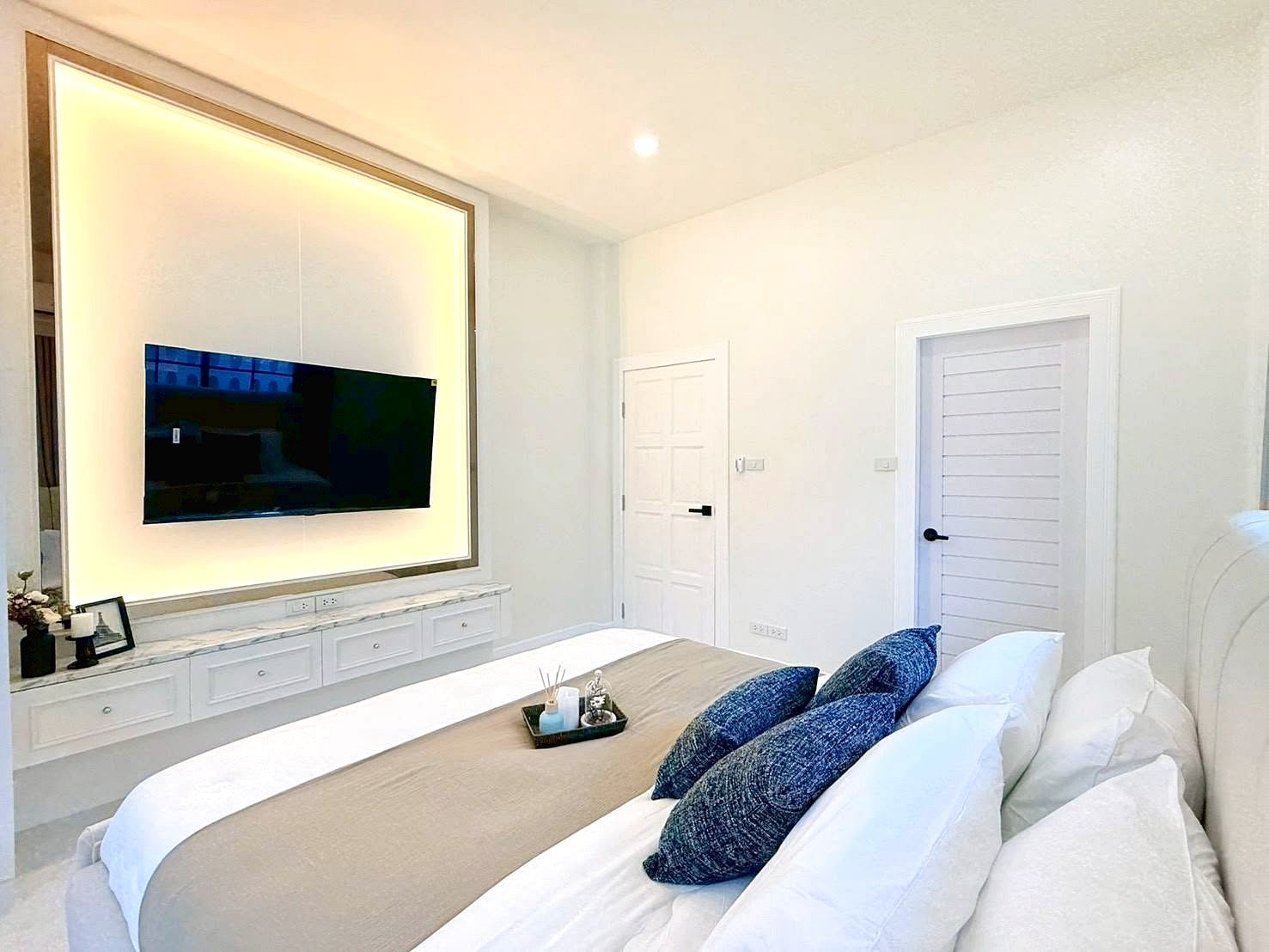 Bright modern bedroom with a wall-mounted flat-screen TV set in a glowing framed inset above a white marble console and drawers, overlooking a neatly made bed with blue accent pillows.