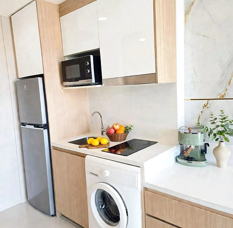 Modern kitchen with light wood cabinets, stainless fridge, microwave, washing machine, sink, and a fruit basket on the counter of a bright space.