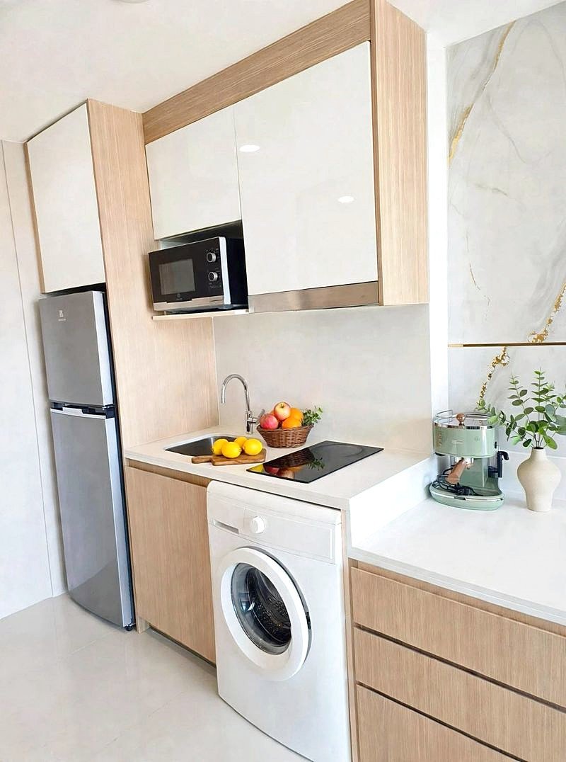 Modern kitchen with light wood cabinets, stainless fridge, microwave, washing machine, sink, and a fruit basket on the counter of a bright space.
