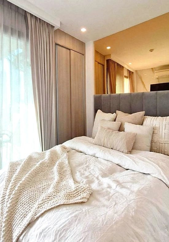 Cozy modern bedroom with a plush gray headboard, cream bedding, and stacked pillows by a window with sheer curtains.