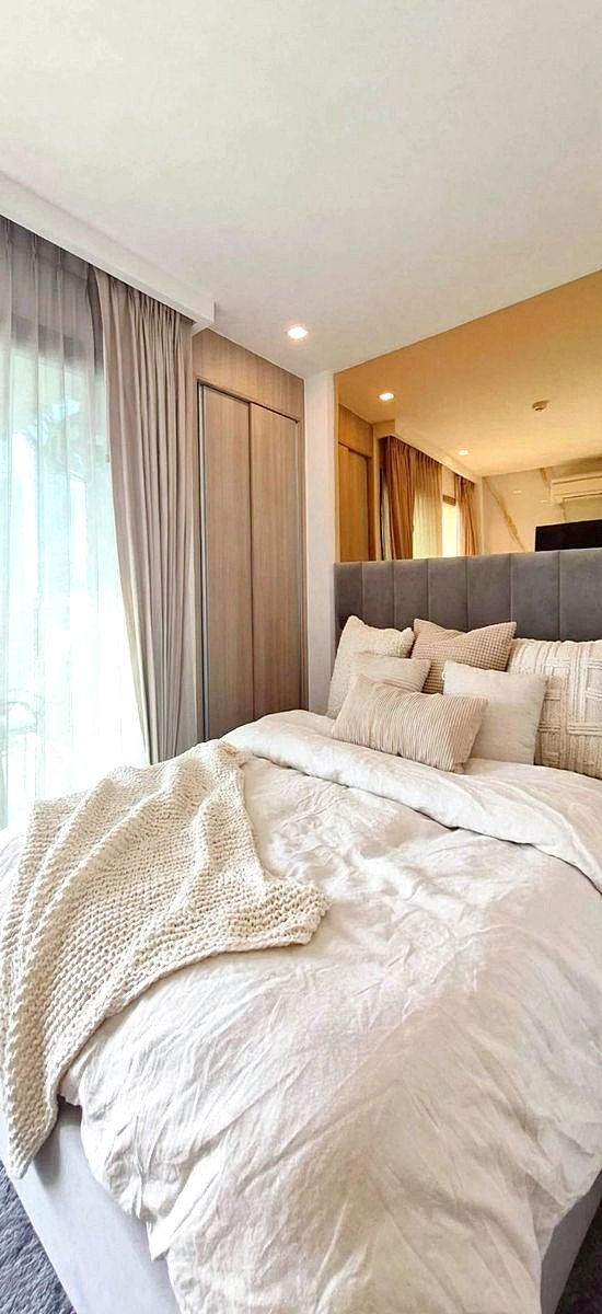 Cozy modern bedroom with a plush gray headboard, cream bedding, and stacked pillows by a window with sheer curtains.