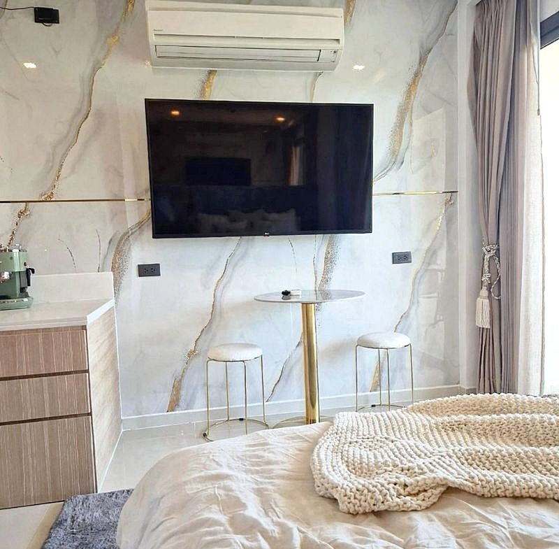 Modern hotel bedroom with a marble feature wall, wall-mounted TV, and a ceiling air conditioner; a small round table with two stools sits by the bed.