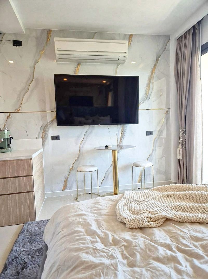 Modern hotel bedroom with a marble feature wall, wall-mounted TV, and a ceiling air conditioner; a small round table with two stools sits by the bed.