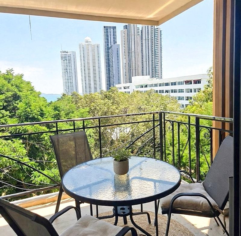 Balcony seating area with a round glass table, small plant, and four chairs overlooking tall city buildings and trees.