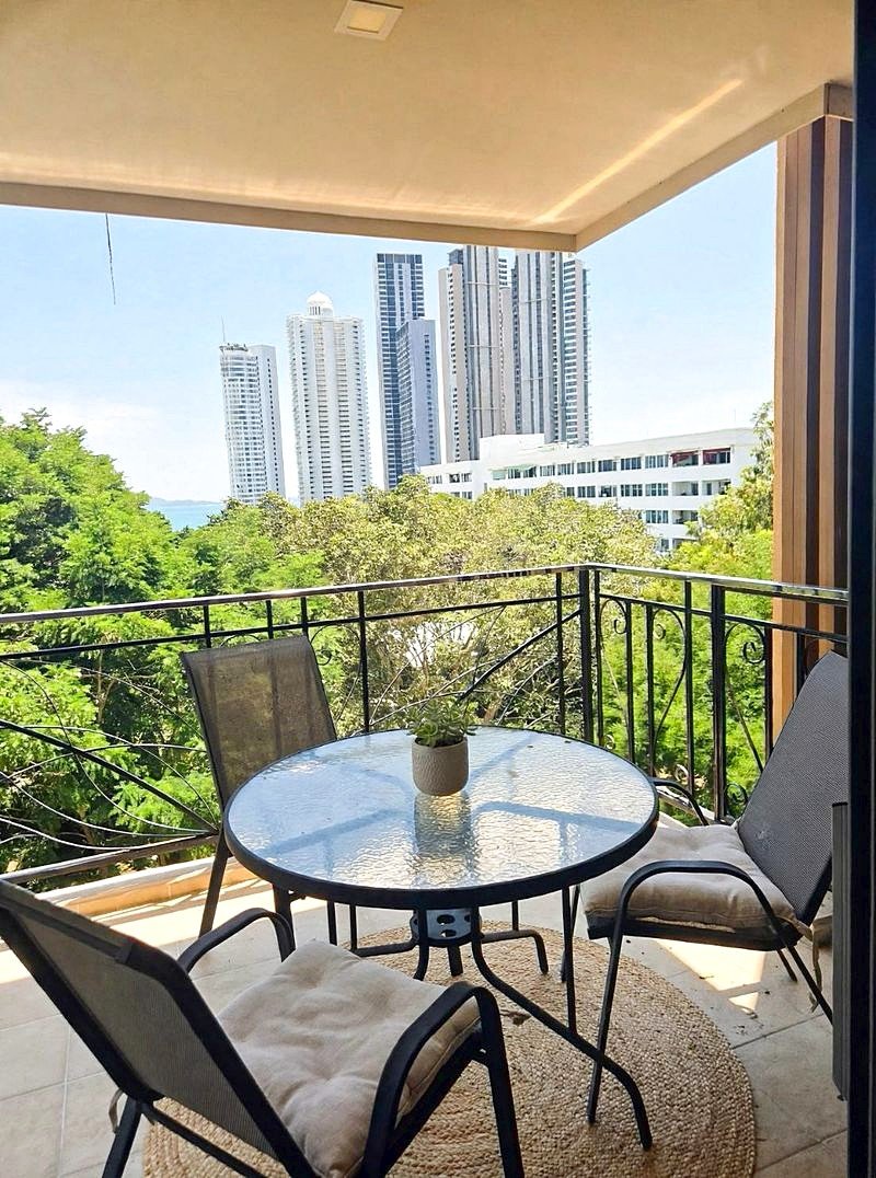 Balcony seating area with a round glass table, small plant, and four chairs overlooking tall city buildings and trees.