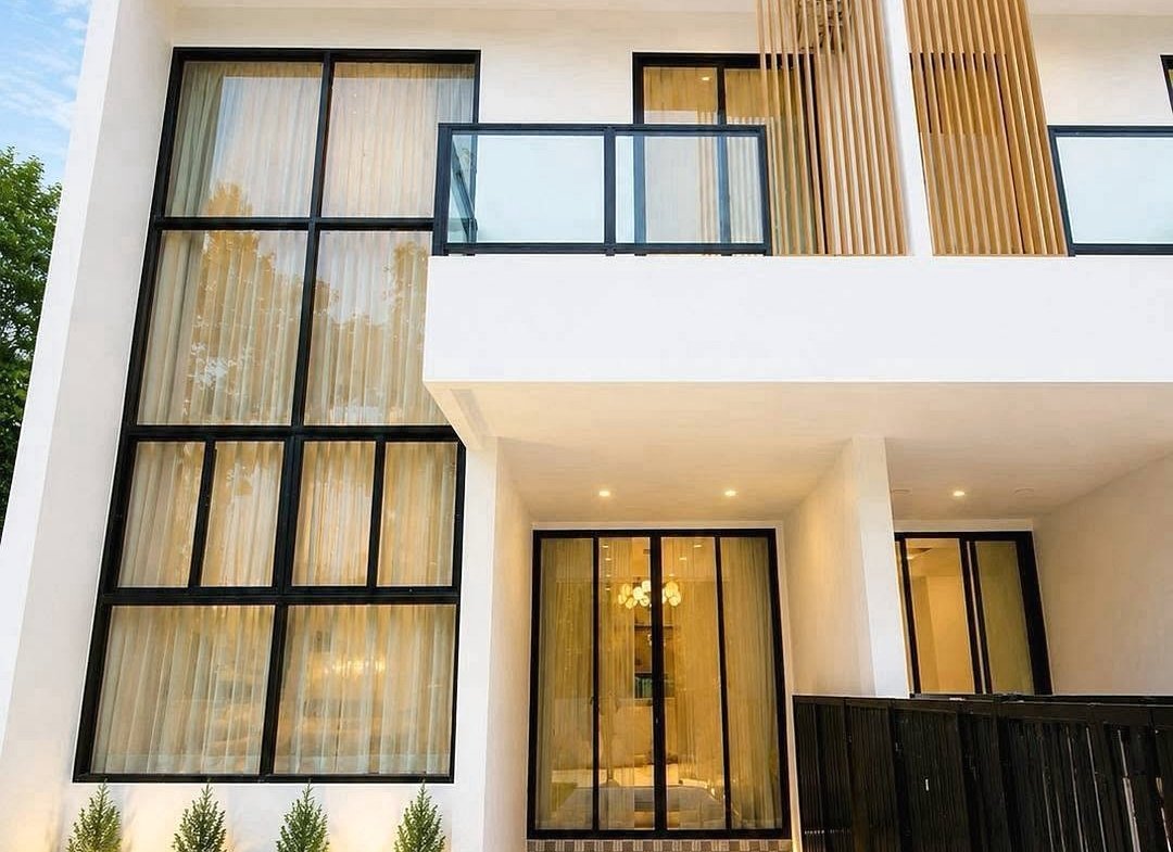 Modern white two-story home with large black-framed windows, a glass balcony, and a tiled entry path under a blue sky.
