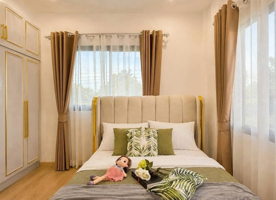 Bedroom with a cream tufted bed, gold trim, and green tropical cushions by sheer, sunlit windows.