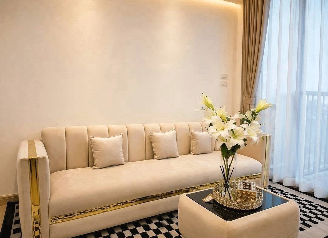 Cream sofa with gold trim and multiple matching pillows in a bright living room setting with a vase of white lilies on a mirrored coffee table.