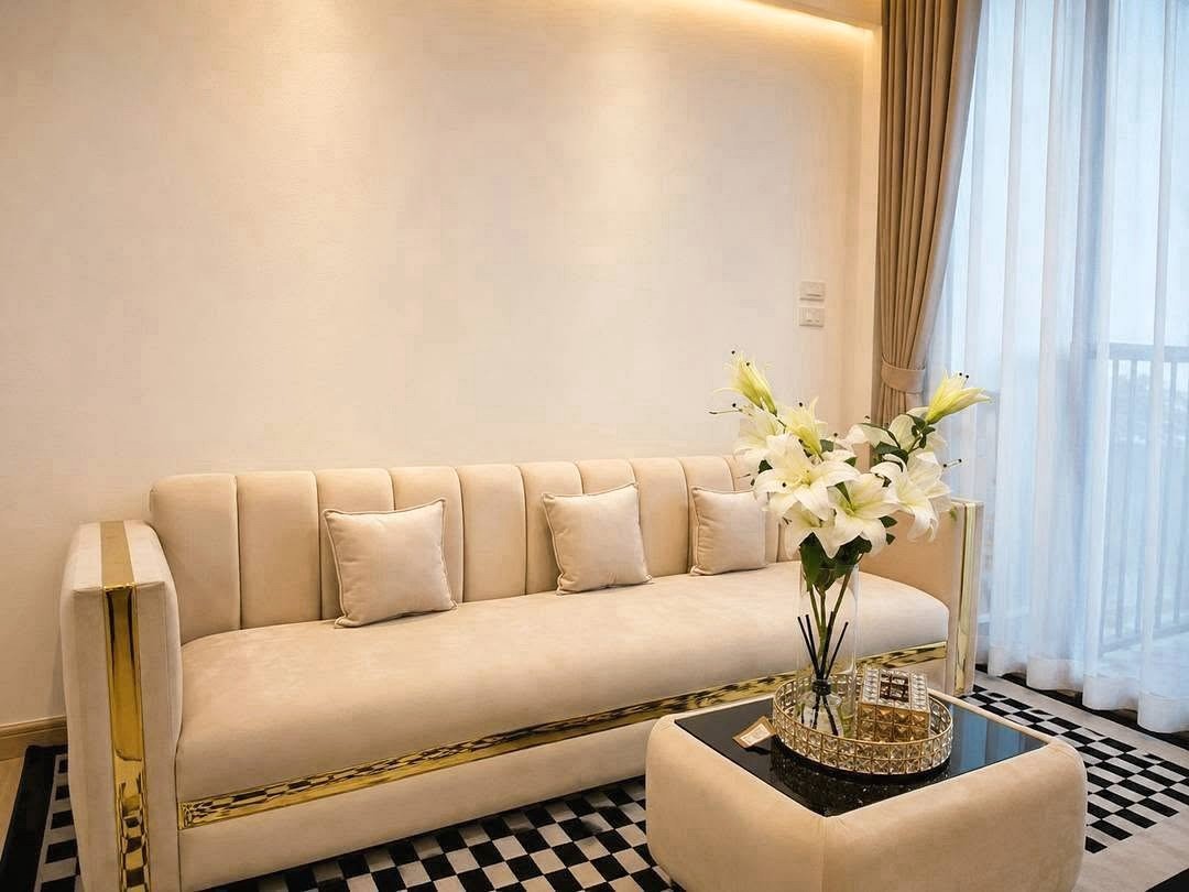 Deperfect_Village_a1_Rdl365_15 Cream sofa with gold trim and multiple matching pillows in a bright living room setting with a vase of white lilies on a mirrored coffee table.