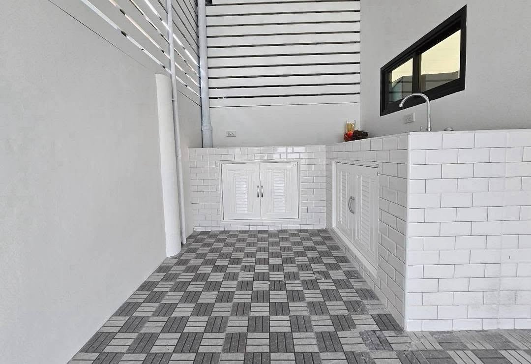 Outdoor kitchenette with white subway-tiled counters, built-in storage cabinets, and a sink under a window; gray checkered floor tiles touch.”