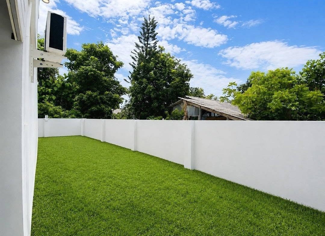 Backyard with a bright green lawn, white privacy fence, and a blue sky with clouds; an outdoor AC unit is mounted on the house wall to the left.