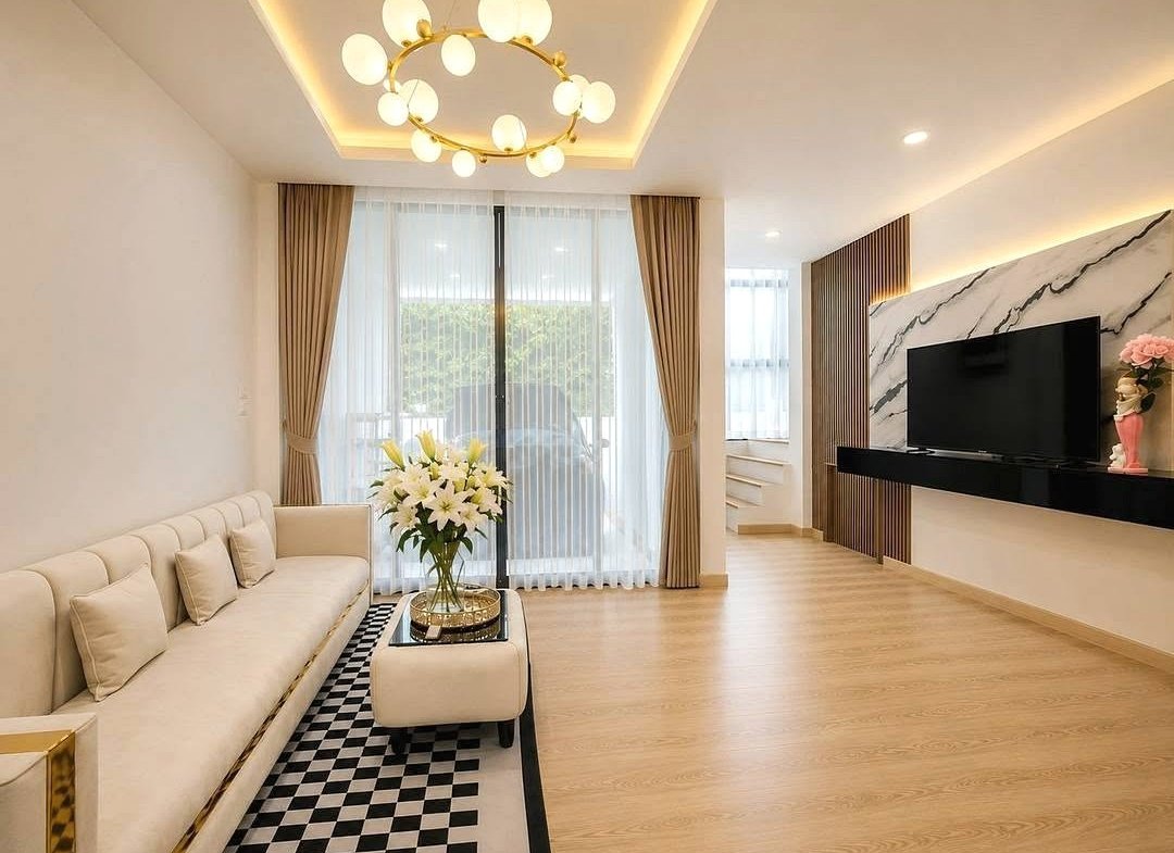 Contemporary living room with a cream sofa, checkered rug, and glass coffee table holding a vase of white lilies; large sliding door with sheer curtains in the background.