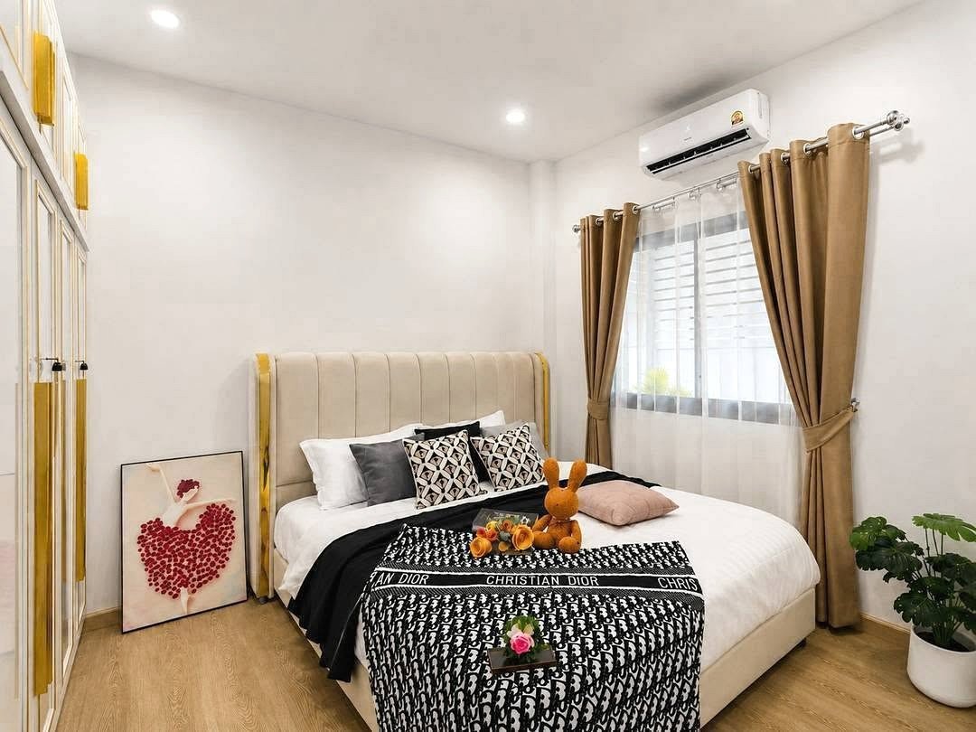 Deperfect_Village_a1_Rdl365_5 Modern bedroom with beige upholstered bed, black-and-white Dior blanket, and decorative pillows; stuffed orange bunny on bed.
