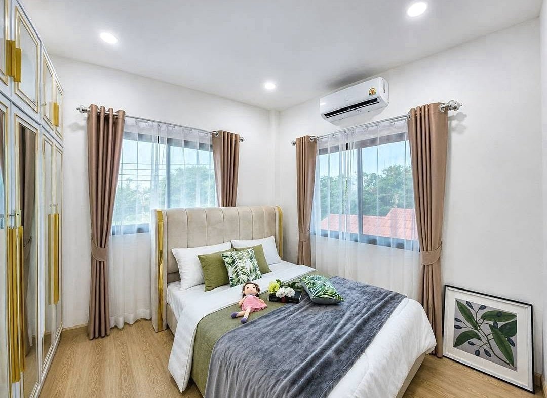 Bright modern bedroom with a beige upholstered bed, green pillows, and a plush doll; windows with sheer white and brown curtains.