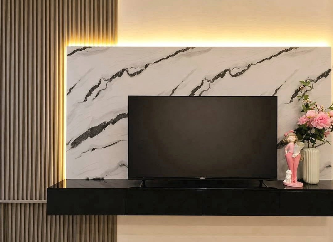 Flat-screen TV mounted on a white marble wall with black veining, glowing backlight, and a black console below. A pink decorative statue and vase with pink flowers sit on the right.