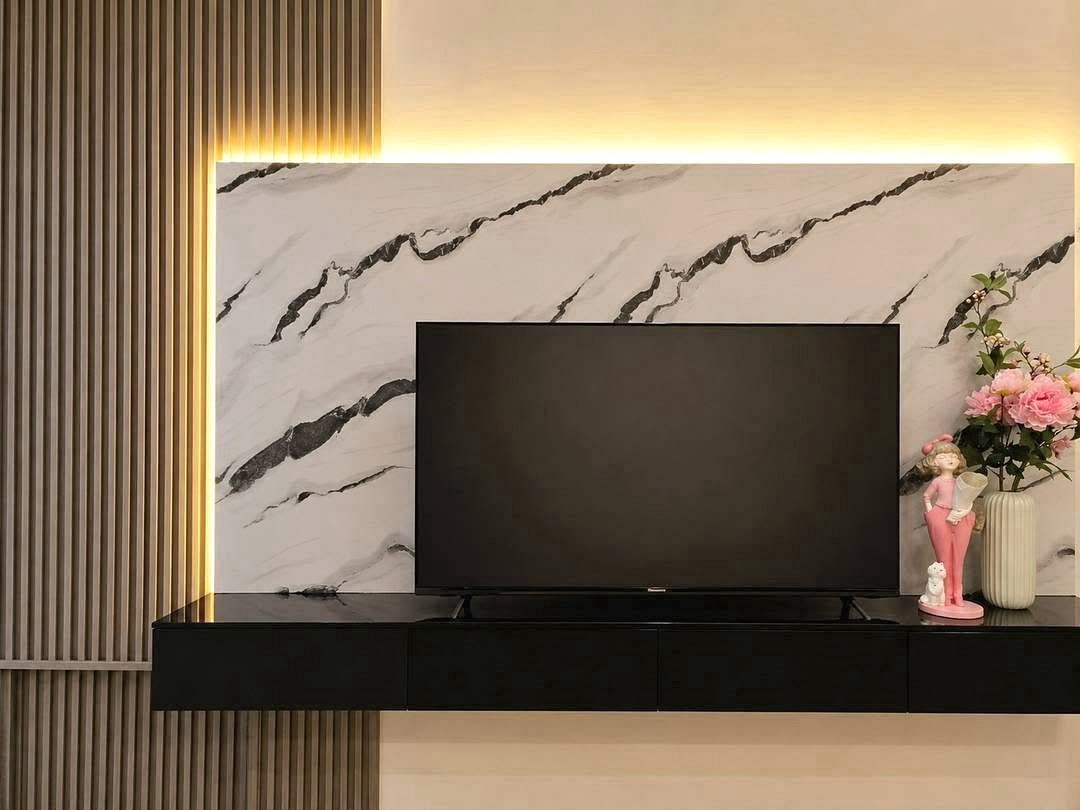 Deperfect_Village_a1_Rdl365_7 Flat-screen TV mounted on a white marble wall with black veining, glowing backlight, and a black console below. A pink decorative statue and vase with pink flowers sit on the right.