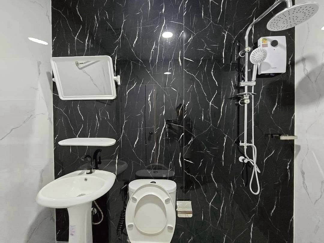 Deperfect_Village_a1_Rdl365_8 Compact bathroom with a white toilet, pedestal sink, and wall-mounted mirror on black marble walls, plus a modern shower system on the right.