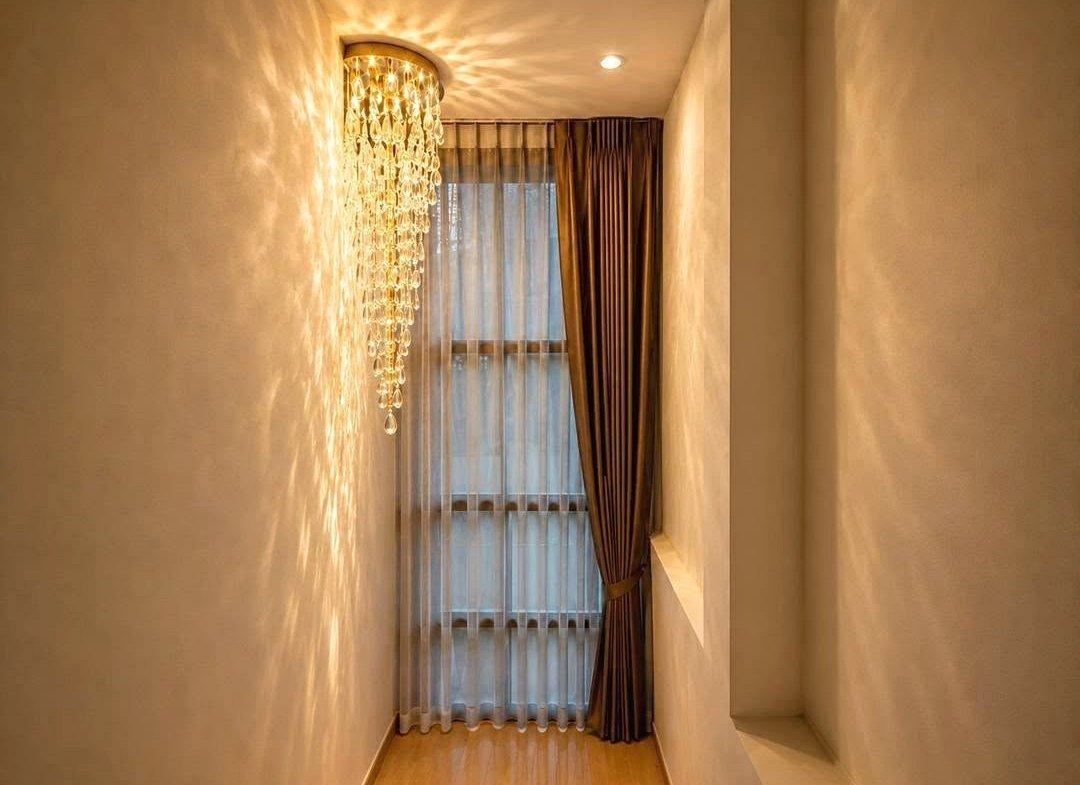 Narrow hallway with a crystal chandelier casting light on pale walls, sheer white curtains and brown drapes over a tall window.