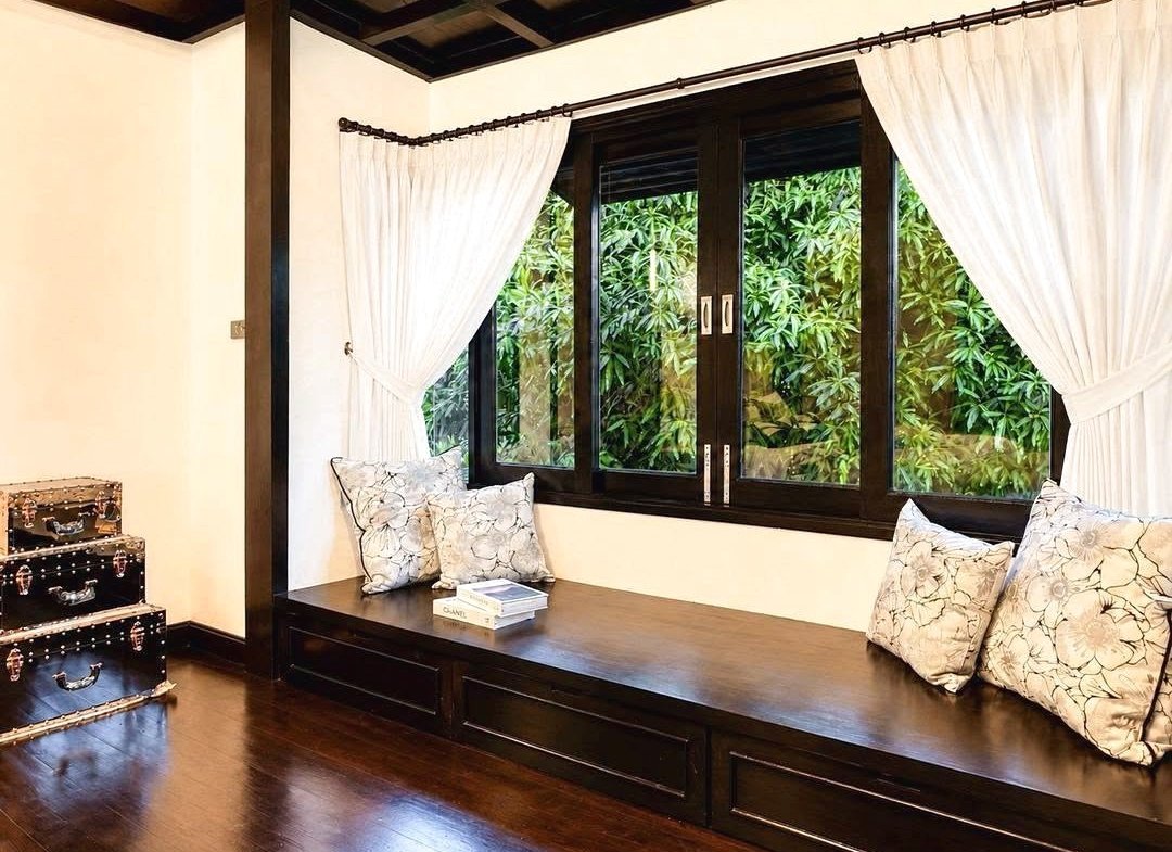 Dark wood window seat with floral cushions and white curtains, overlooking green bushes; vintage suitcases stacked to the left.