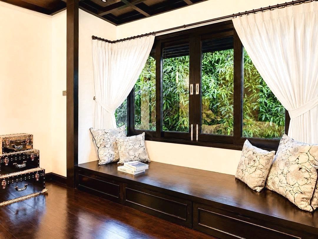 Dharawadi_Poolvilla_Rent_Rdl353_12 Dark wood window seat with floral cushions and white curtains, overlooking green bushes; vintage suitcases stacked to the left.
