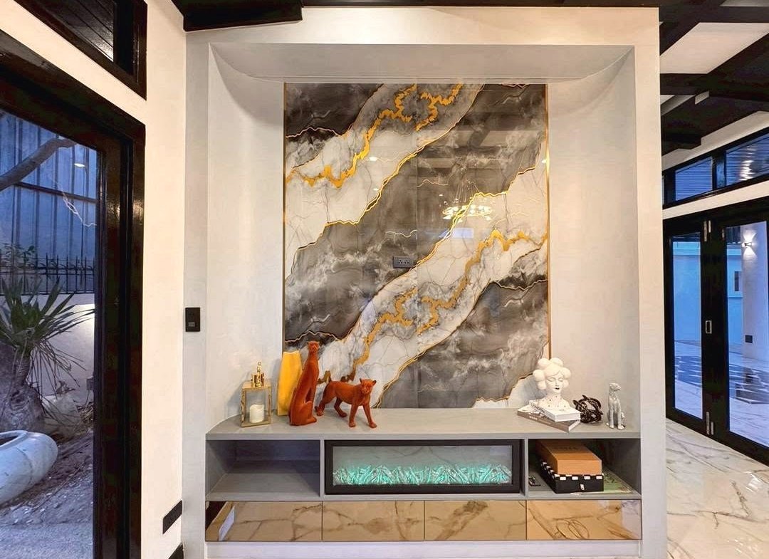 A modern entryway with a large marble wall panel featuring gold veining above a gray console cabinet displaying orange animal sculptures, a white bust, books, and decorative objects, with teal backlighting in the bottom drawer.