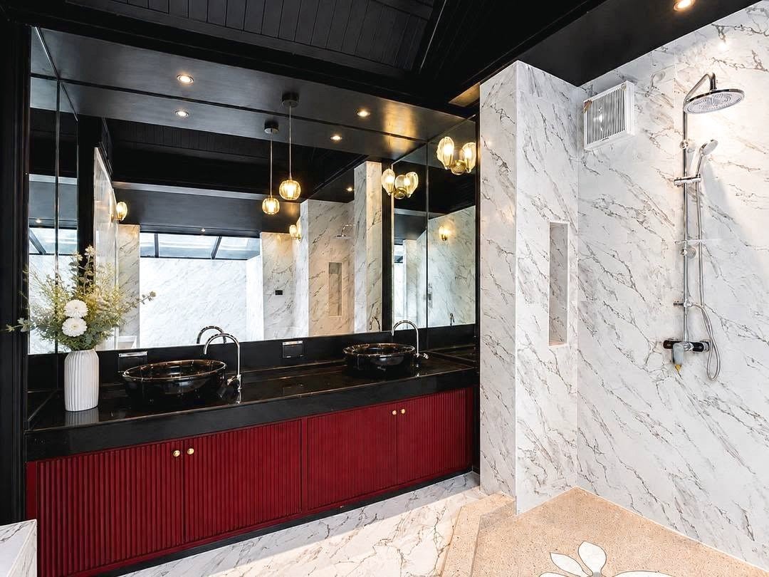 Dharawadi_Poolvilla_Rent_Rdl353_14 Luxurious marble bathroom with a dual-sink vanity, black countertop, and red cabinetry next to a glass-enclosed shower.