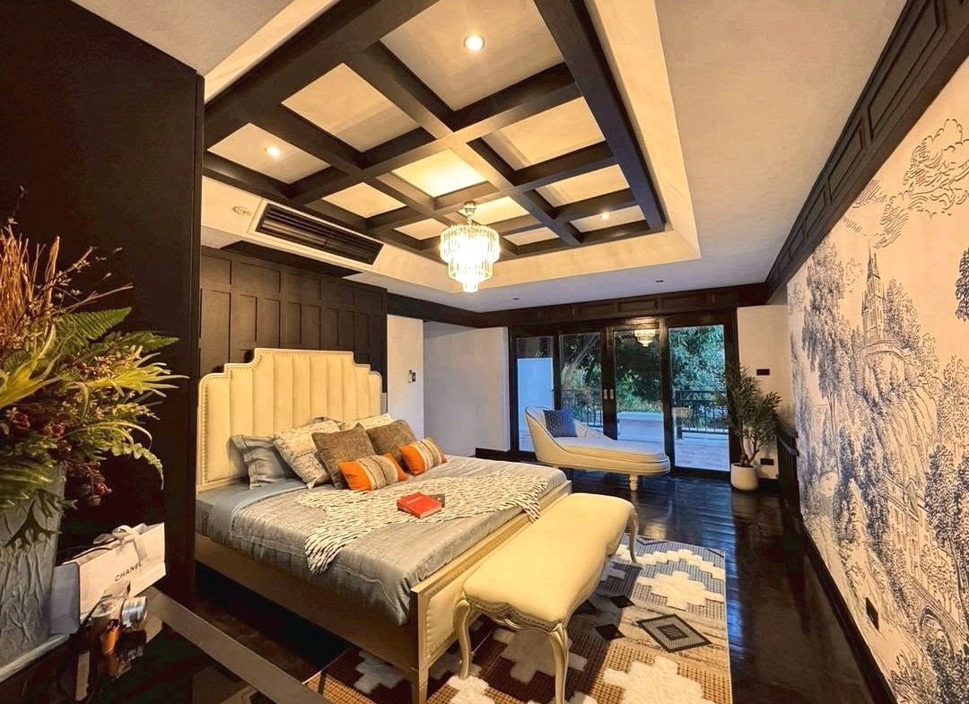 Luxurious bedroom with a tufted headboard bed, chandelier, and woven bench at foot, dark wood ceiling beams, and a mural wall on the right.
