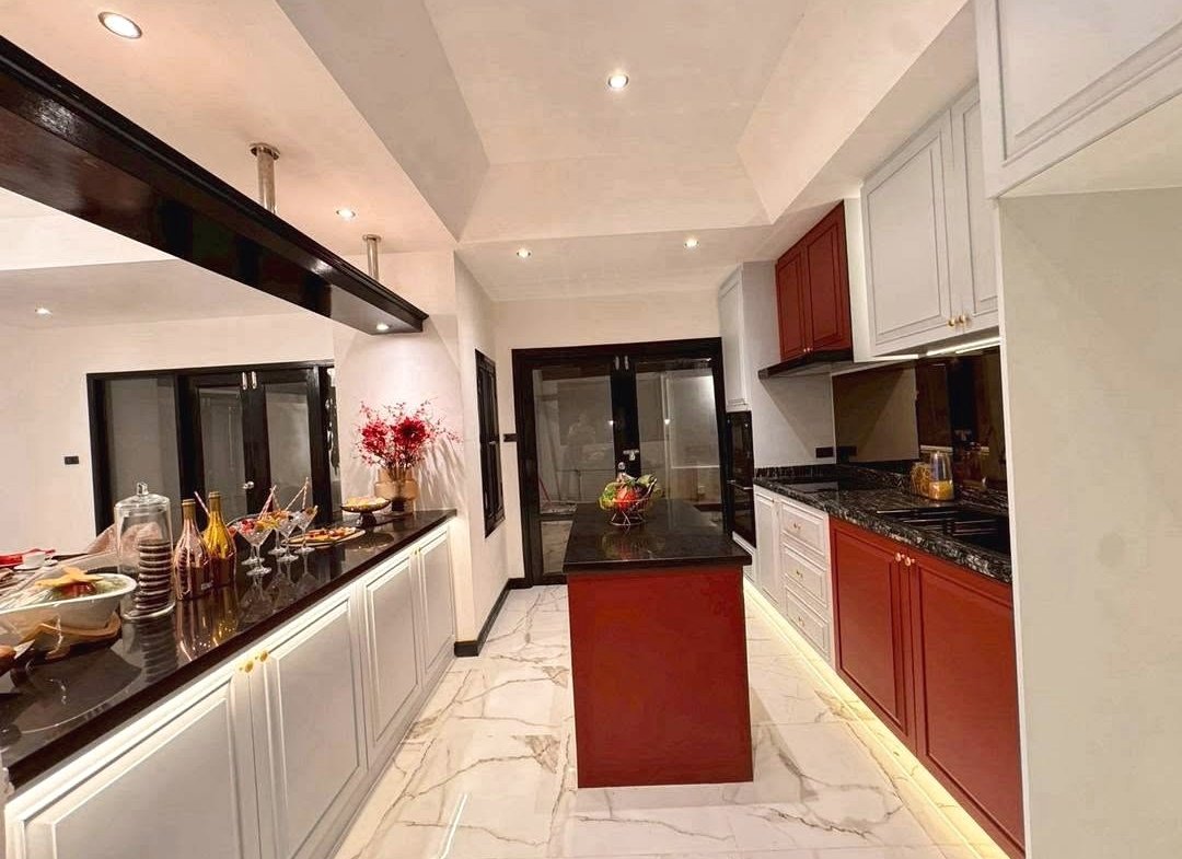Modern L-shaped kitchen with white upper cabinets, red lower cabinets, and a central island on a marble floor; warm under-cabinet lighting and a beverage setup on the left counter.