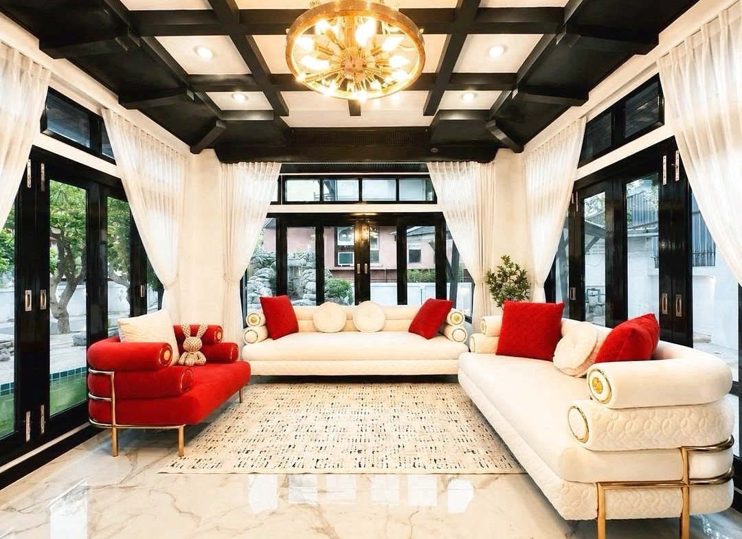 Bright, modern living room with white sofas, red cushions, black ceiling beams, and a gold chandelier at the center.