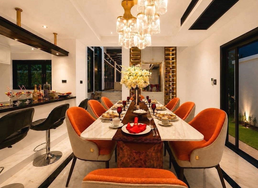 Luxurious dining room with a long marble table, orange upholstered chairs, and a dramatic crystal chandelier set for dinner.