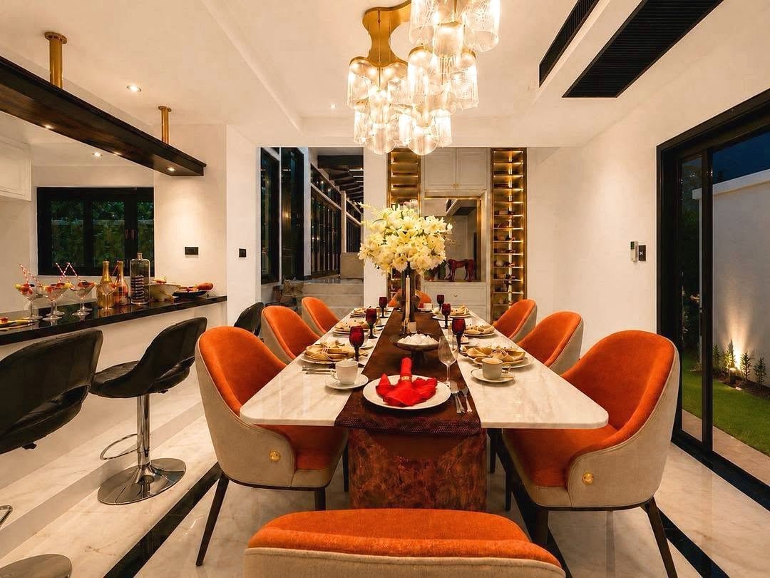 Dharawadi_Poolvilla_Rent_Rdl353_4 Luxurious dining room with a long marble table, orange upholstered chairs, and a dramatic crystal chandelier set for dinner.
