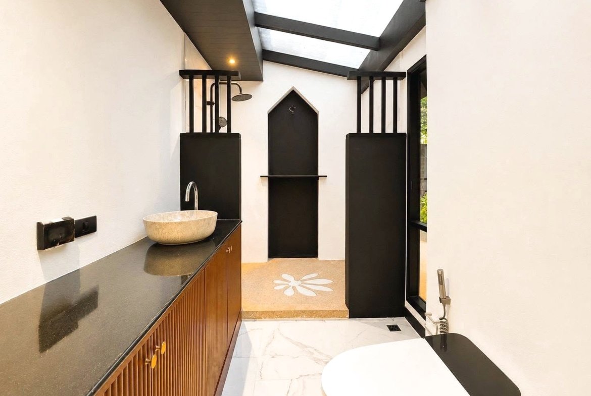 Modern bathroom with a circular stone sink on a dark countertop, wooden cabinet, and a skylight overhead.
