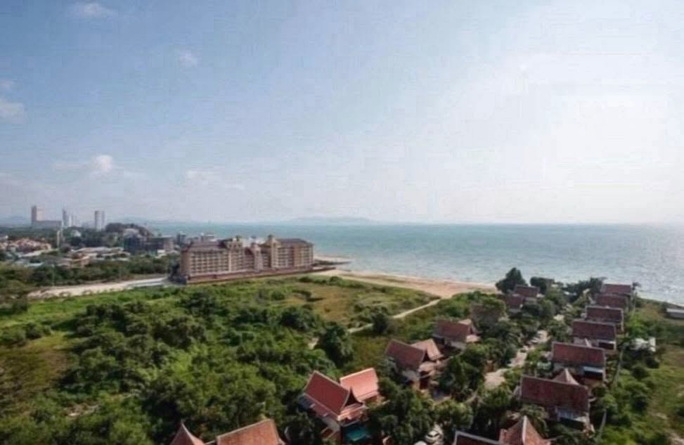 A coastal scene showing a sandy beach, calm sea, and a cluster of red-roofed houses among green vegetation with larger resort buildings in the distance.
