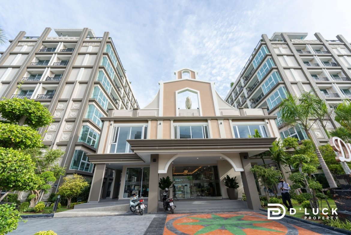Front view of a modern beige apartment complex with two wings, glass balconies, and a central arched entrance with a statue, greenery on sides.