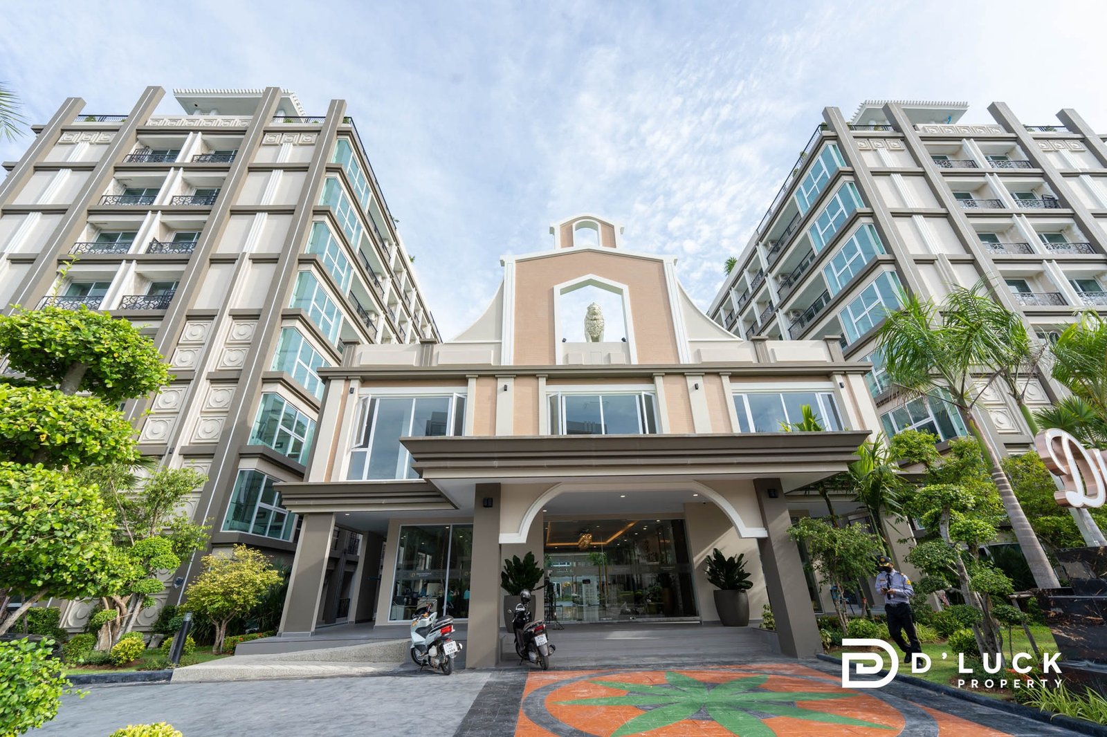 Dusit Grand Park 2 1 Front view of a modern beige apartment complex with two wings, glass balconies, and a central arched entrance with a statue, greenery on sides.