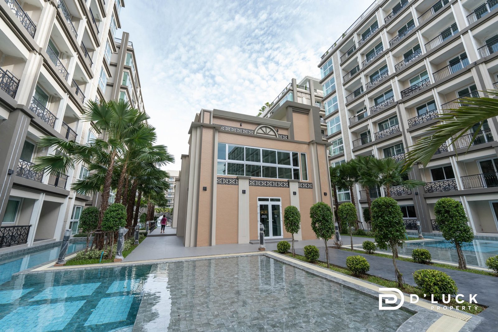 Dusit Grand Park 2 2 Modern apartment courtyard with beige buildings, balconies, and palm trees surrounding a shallow, reflective pool.