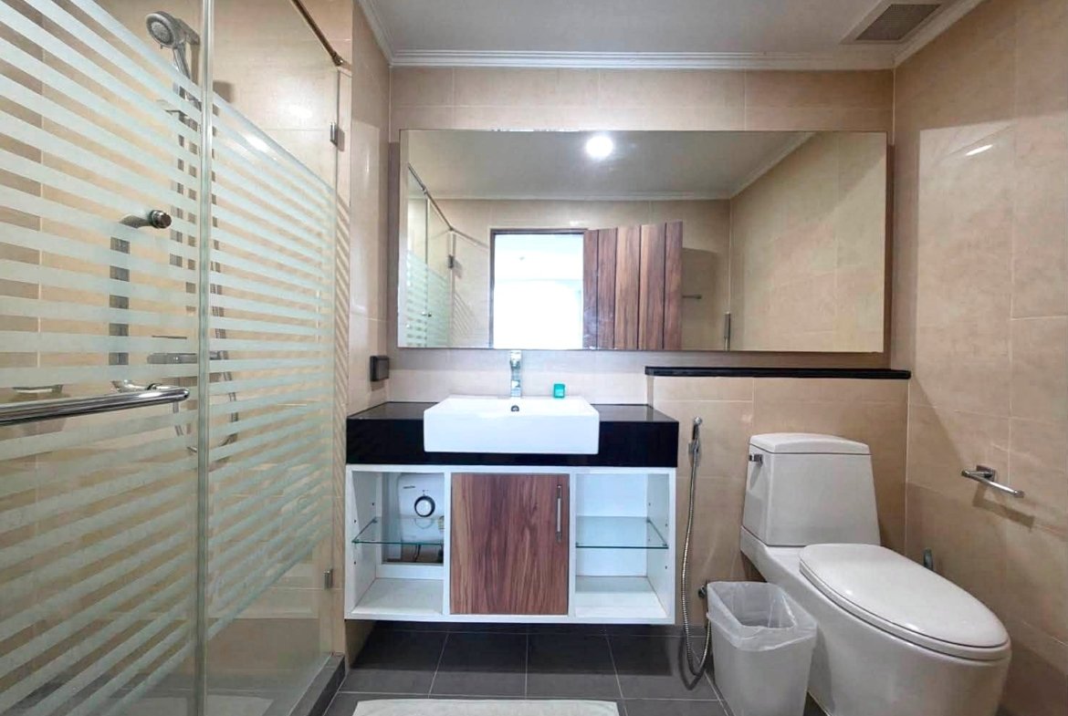 Modern bathroom with a glass shower, white sink on a black countertop, large mirror, toilet, and beige tile walls.